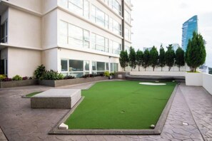 Un putting green en la azotea con un hoyo de golf, rodeado por un muro de piedra bajo, y contiguo a un edificio moderno con grandes ventanales.