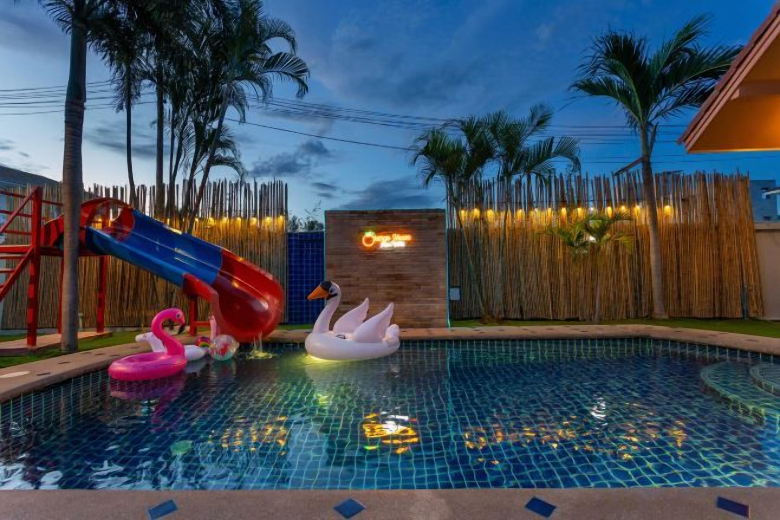 A swimming pool with a slide and inflatable swans, surrounded by palm trees and a wooden fence.