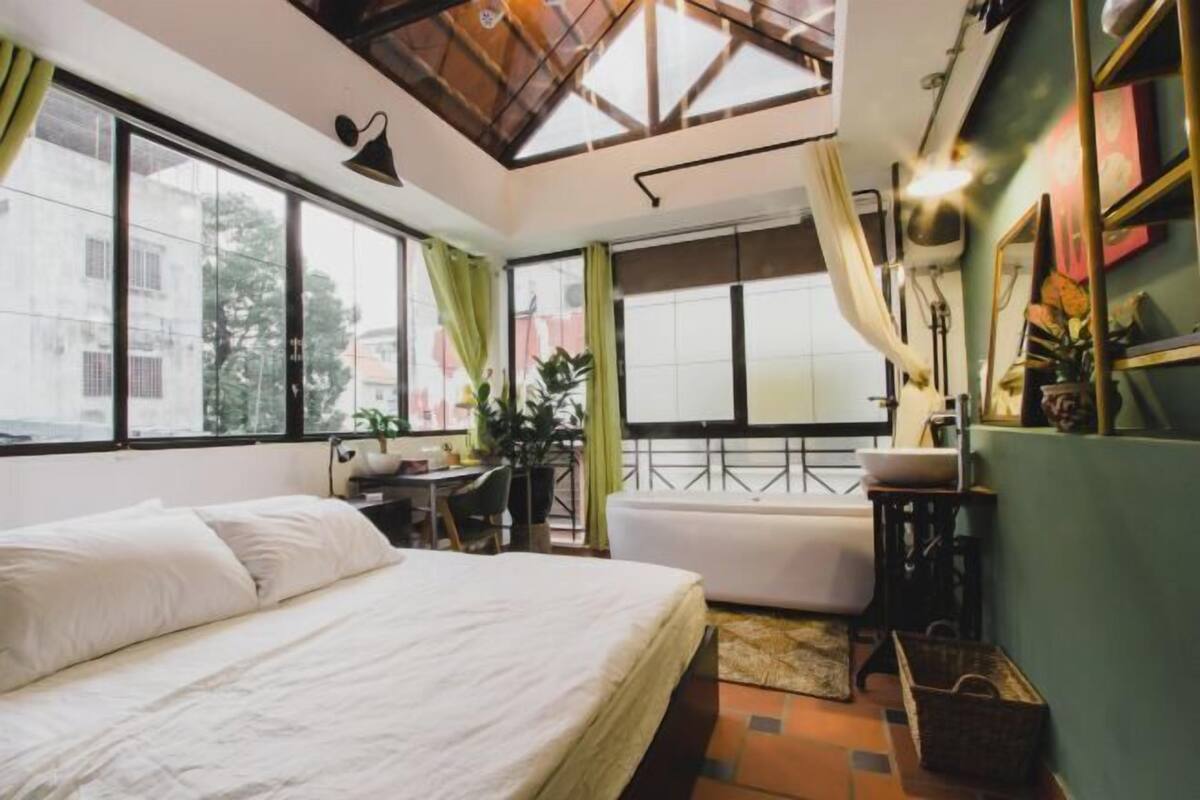 A bedroom with a bed, a desk, a bathtub, and a view of the outside.