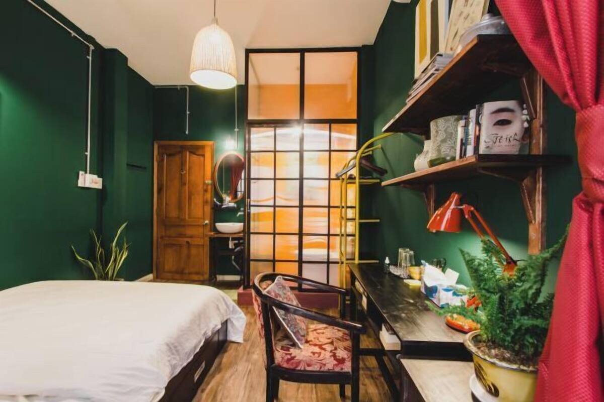 A room with a bed, a desk, a chair, a potted plant, and a shelf with books and a lamp.