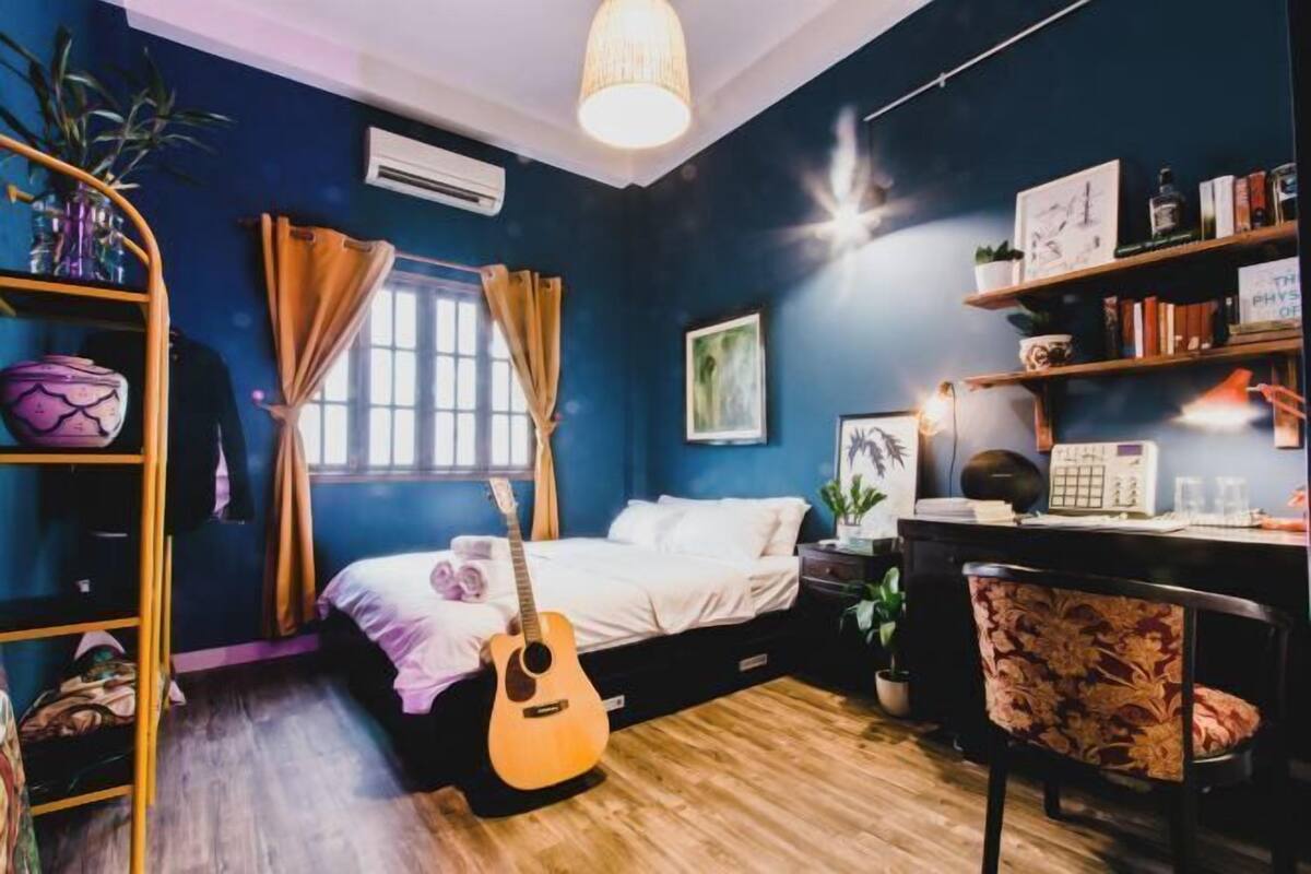 A bedroom with a bed, guitar, desk, chair, and shelves with books and plants.