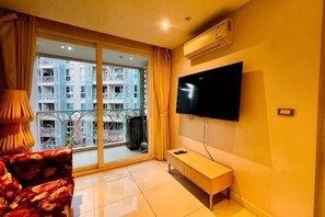 A living room with a TV, a sofa, a bench, and a large window with a view of a building. - Large Caribbean 1BR Condo near Jomtien Bus Station (Pattaya)