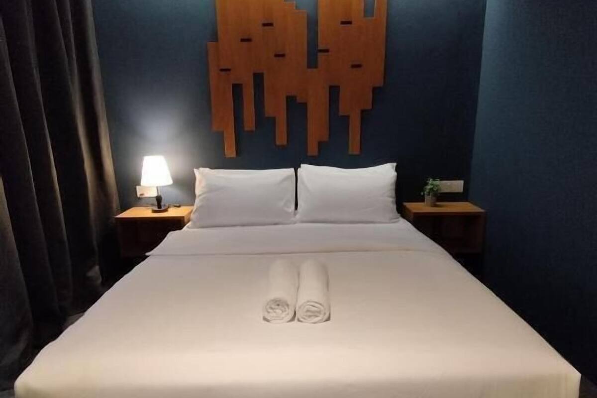 A neatly made bed with white linens and rolled towels, a wooden headboard with a unique design, a bedside lamp, and a small table with a plant.