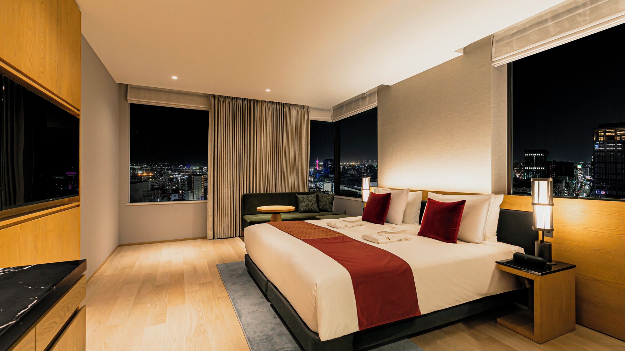A modern hotel room with a large bed, a desk, and a view of a cityscape at night.