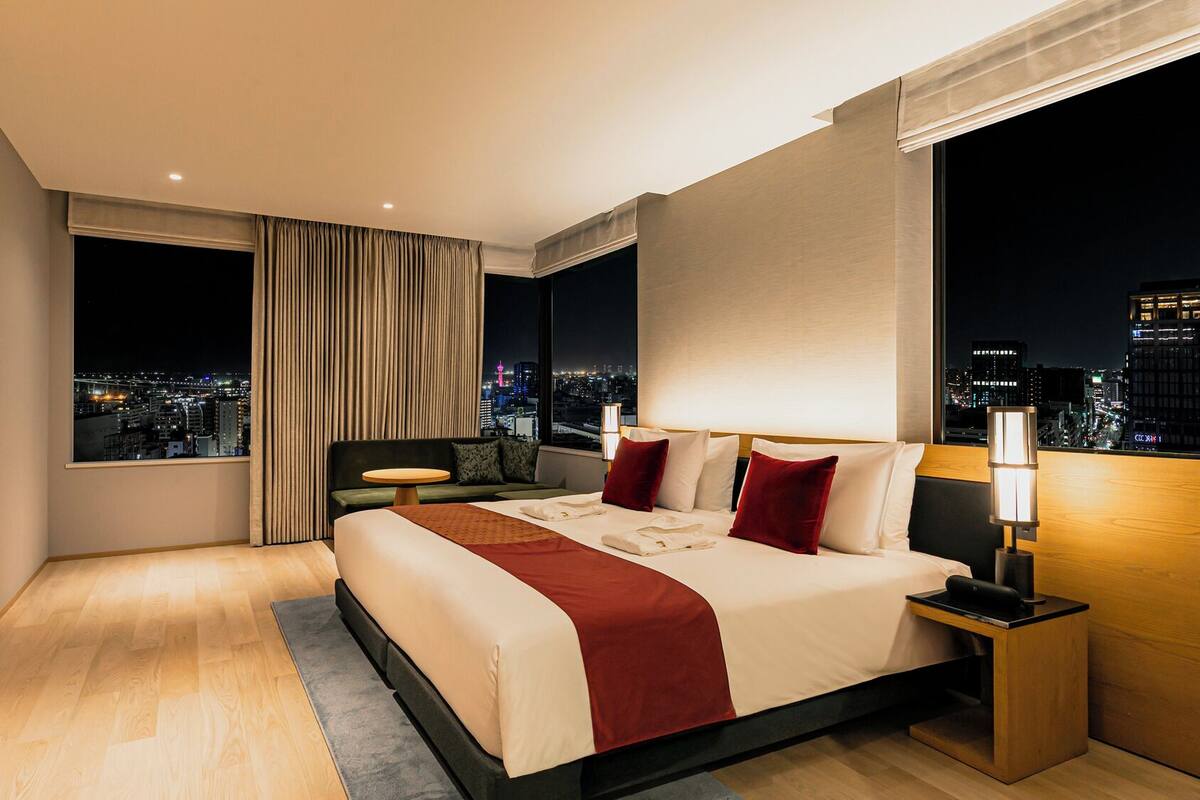 A modern hotel room with a large bed, a desk, and a view of a cityscape at night.