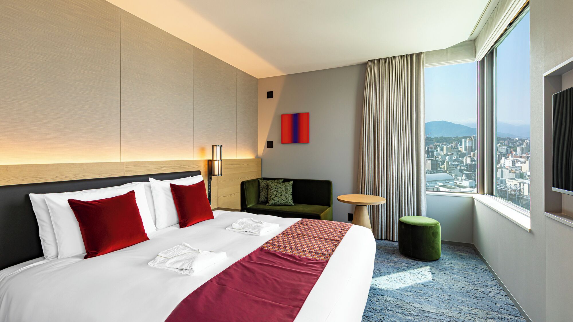 A hotel room with a bed, a sofa, a small table, a TV, and a window with a city view.