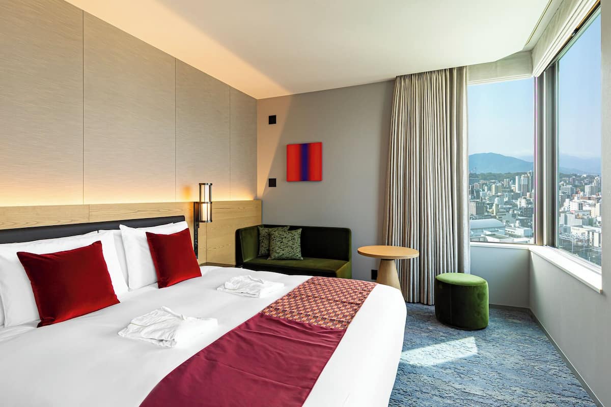 A hotel room with a bed, a sofa, a small table, a TV, and a window with a city view.