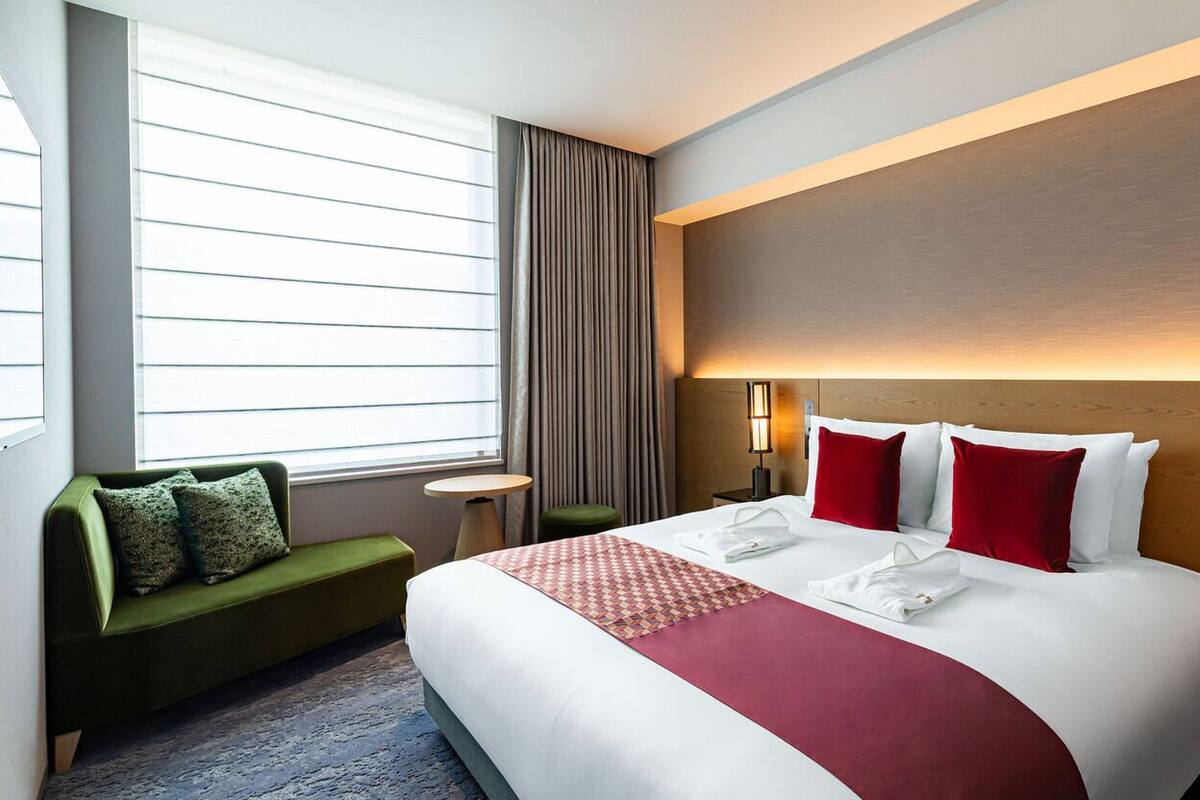 A hotel room with a large bed, a green sofa, a small round table, and a window with blinds.