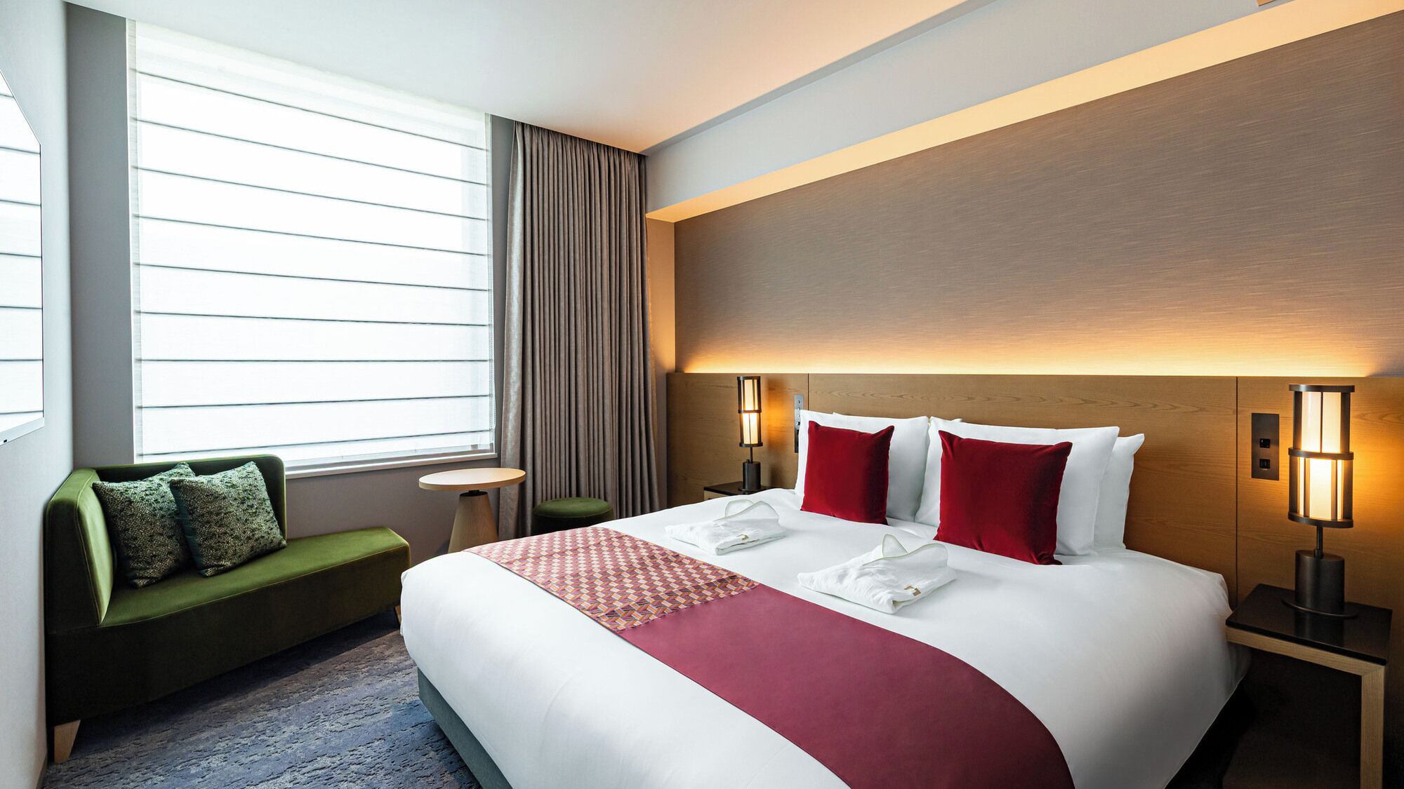 A hotel room with a large bed, a green sofa, a small table, and a window with blinds.