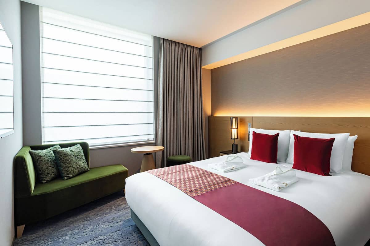 A hotel room with a large bed, a green sofa, a small table, and a window with blinds.