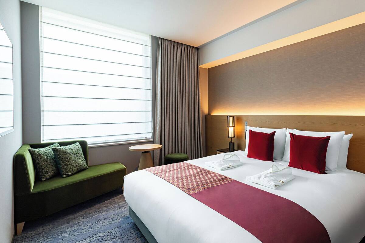A hotel room with a large bed, a green sofa, a small table, and a window with blinds.