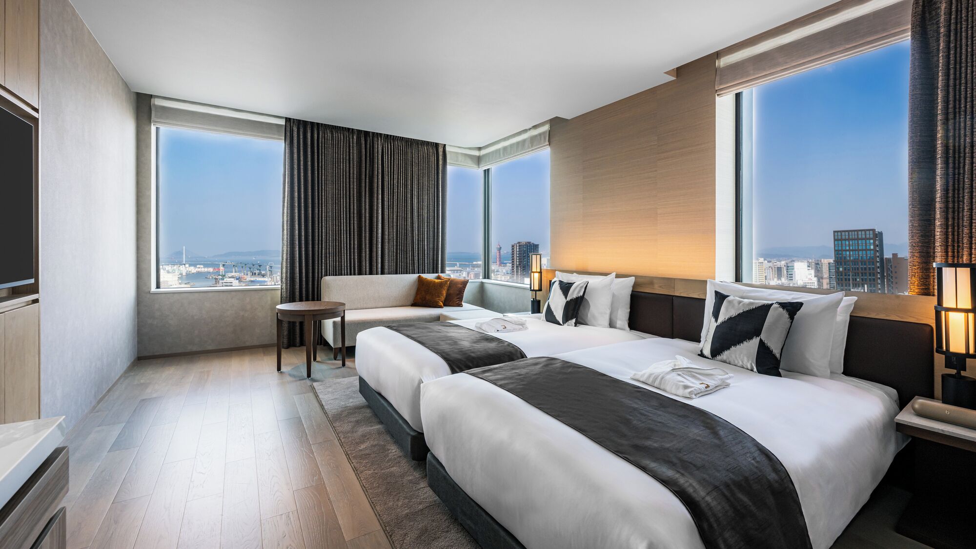 A hotel room with two beds, a sofa, a small table, and large windows offering a city view.