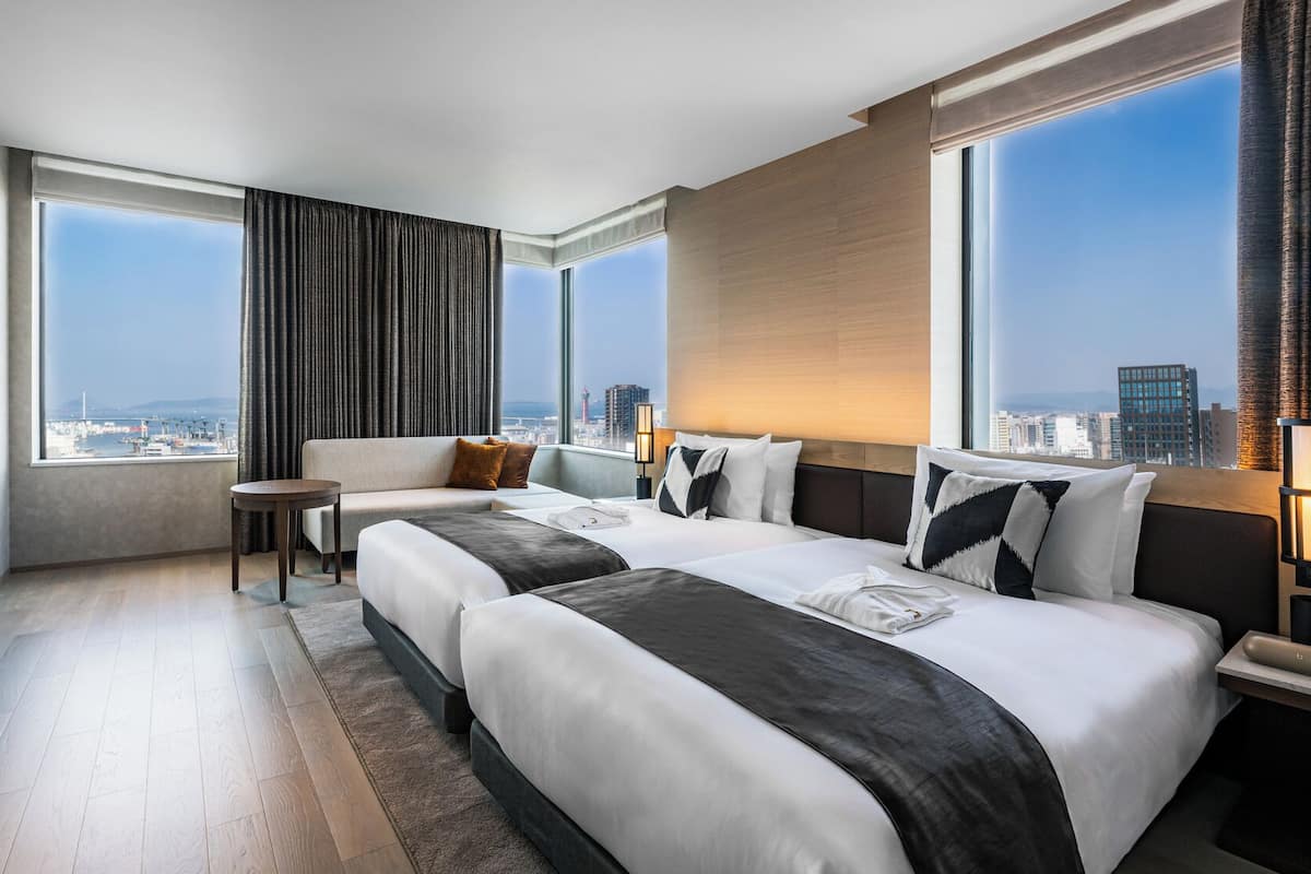 A hotel room with two beds, a sofa, a small table, and large windows offering a city view.