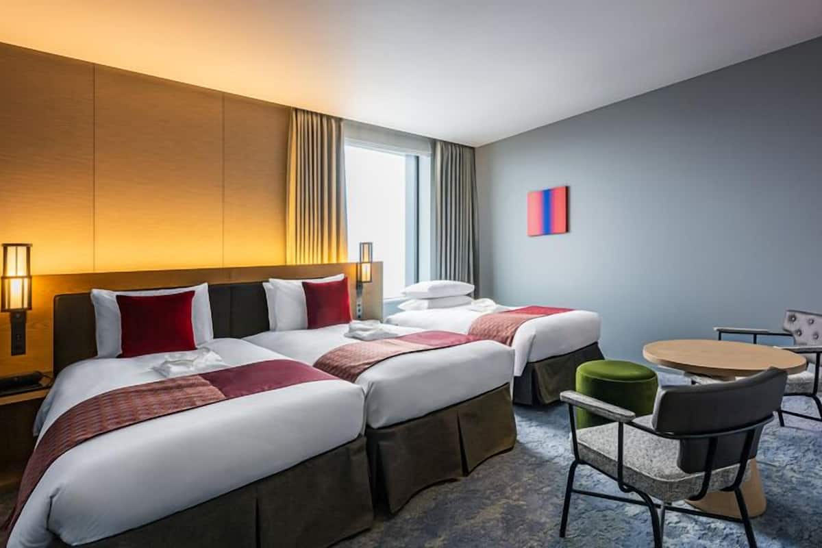 A hotel room with two beds, a small round table, a chair, and a lamp.