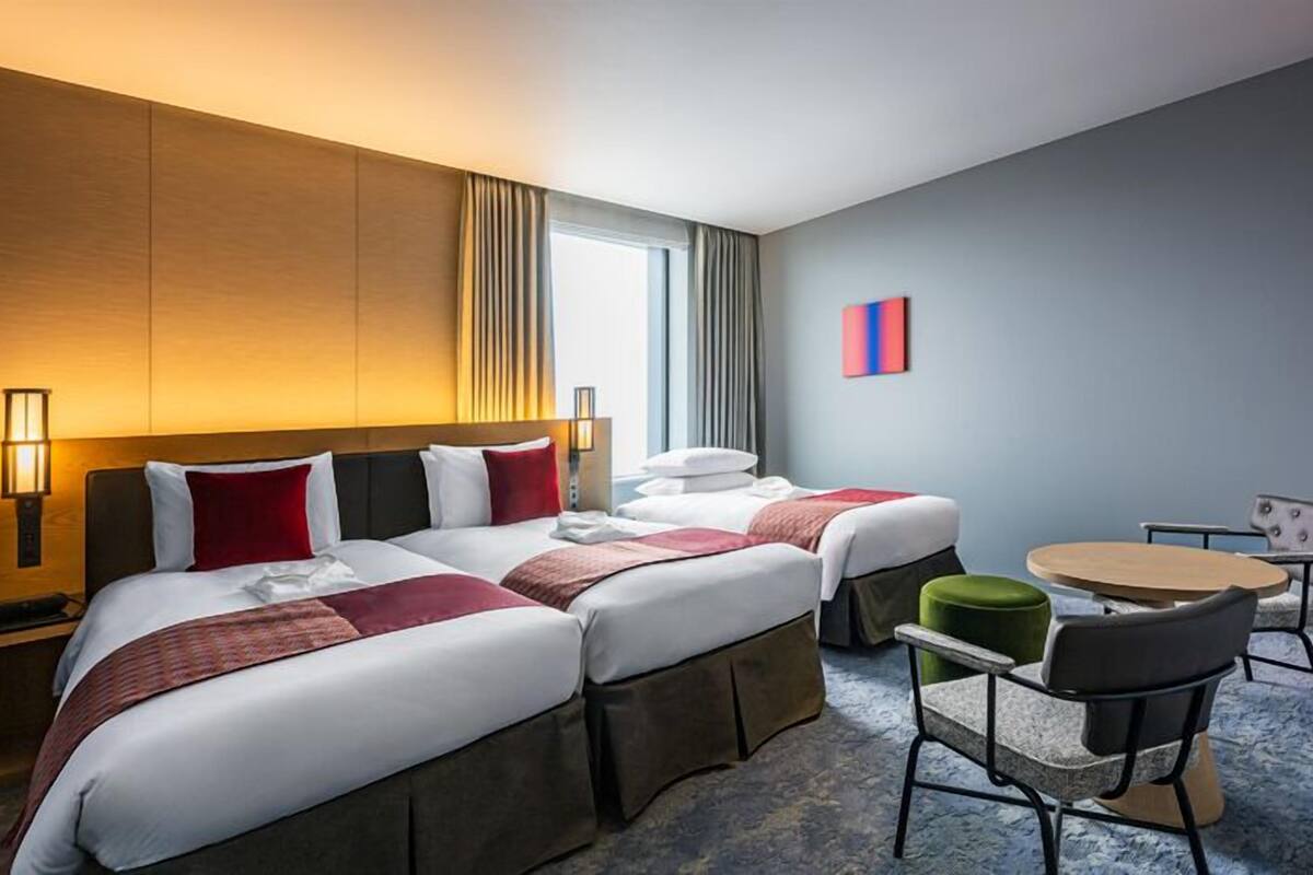 A hotel room with two beds, a small round table, a chair, and a lamp.