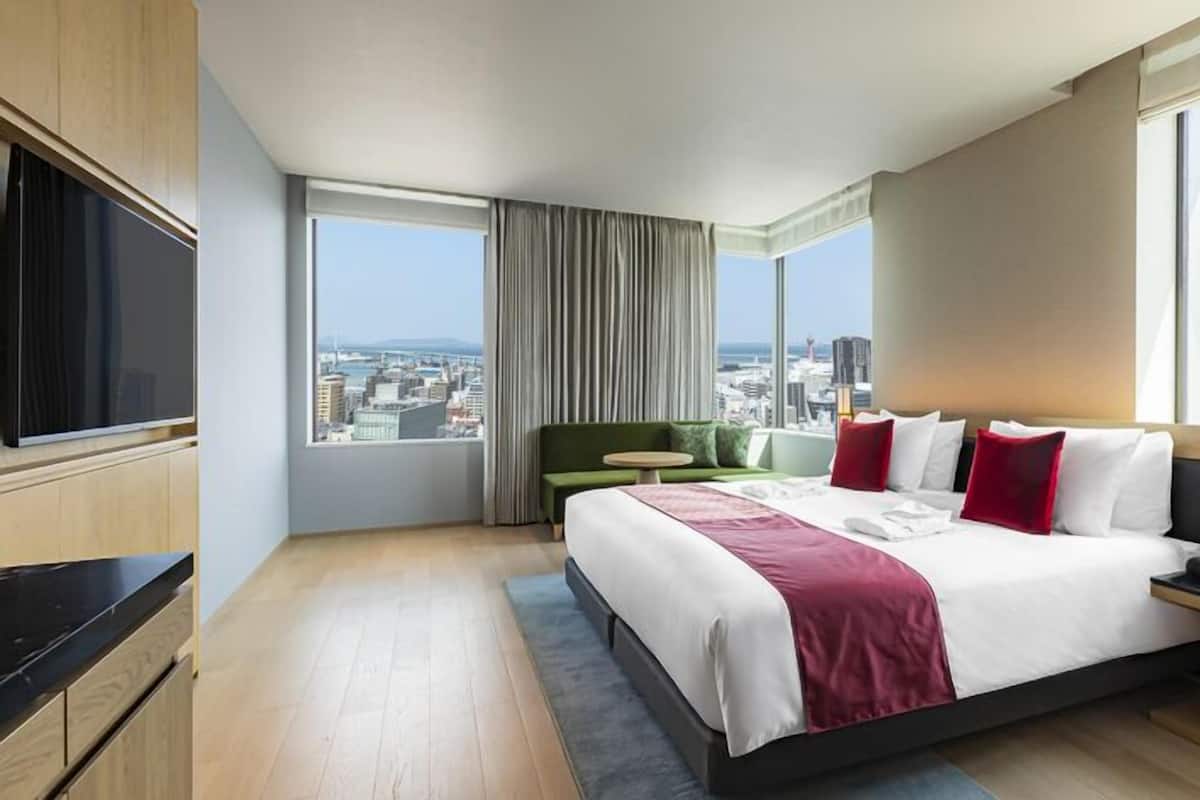 A hotel room with a large bed, a TV, a city view, and a green chair.