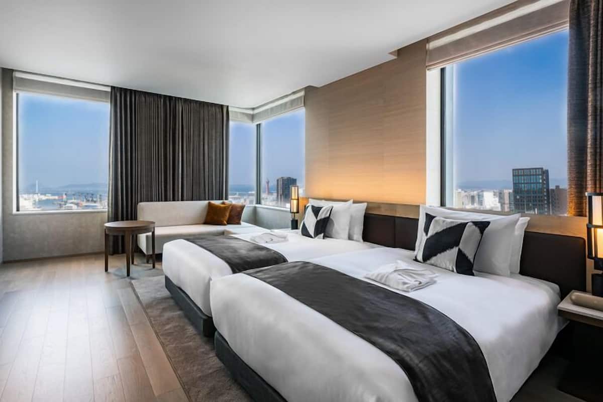 A hotel room with two beds, a sofa, a small table, and large windows offering a city view.