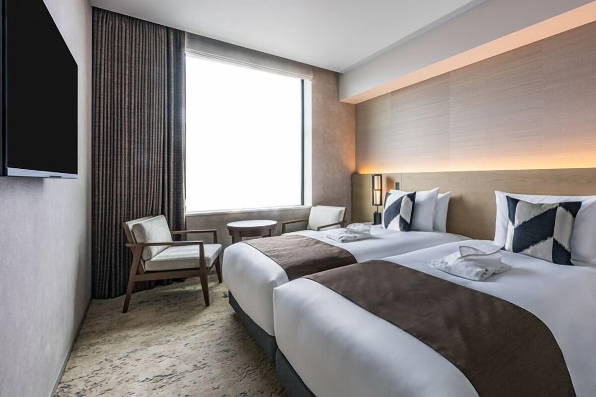 A hotel room with two beds, a TV, a window with curtains, a desk, and a chair.