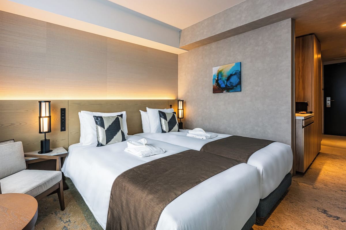 A hotel room with two beds, a desk, a chair, and a painting on the wall.