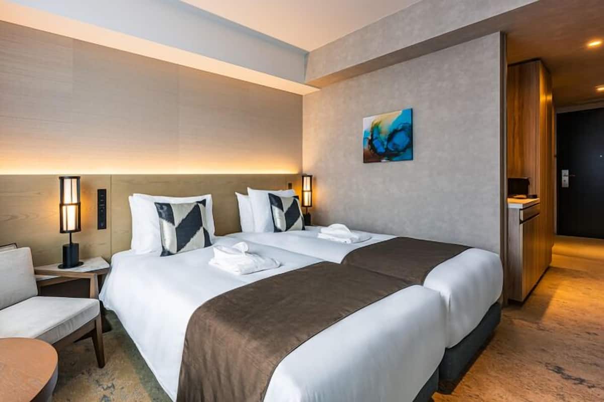 A hotel room with two beds, a chair, a table, and a painting on the wall.