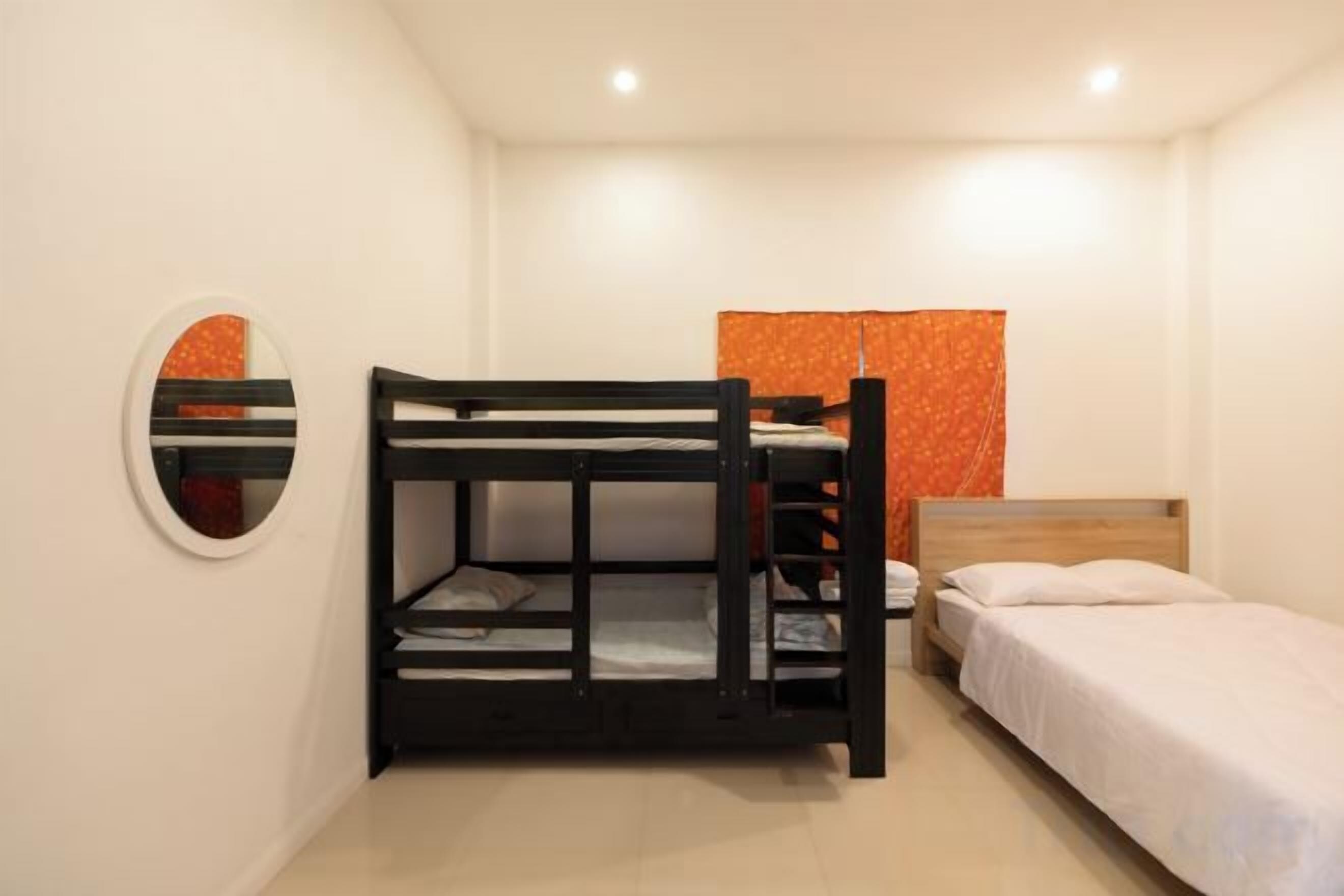 A bunk bed with a lower bunk and a single bed, a round mirror on the wall, and a window with orange curtains.