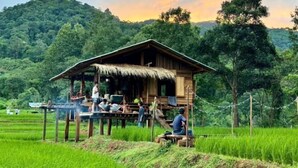 A wooden structure with a thatched roof, surrounded by lush green fields and hills in the background. - Hae o bi (rice terraces) (Samoeng)