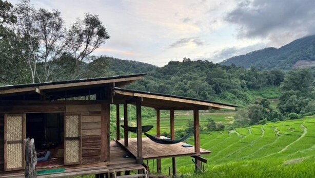A wooden cabin with a porch and a hammock, surrounded by lush green rice paddies and mountains in the background. - Hae o bi (rice terraces) (Samoeng)