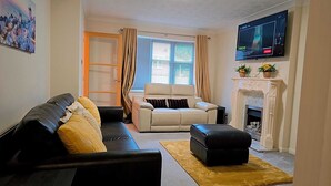 A living room with a black sofa, a beige armchair, a black ottoman, a fireplace, and a television mounted on the wall.