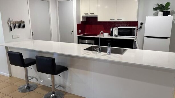 A modern kitchen with white cabinets, a red backsplash, a microwave, and a refrigerator. There are two black bar stools at the island. - Family retreat in Caboolture big house (Brisbane)