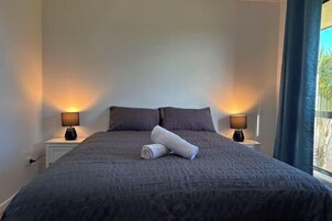 Una cama bien tendida con una toalla enrollada, dos lámparas de noche y una ventana con cortinas.