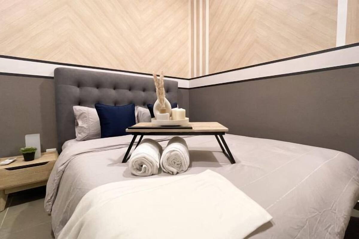 Una habitación de hotel moderna con un cabecero capitonado, una cama bien hecha con ropa blanca, una mesita de noche pequeña con una planta y un tocador de madera.
