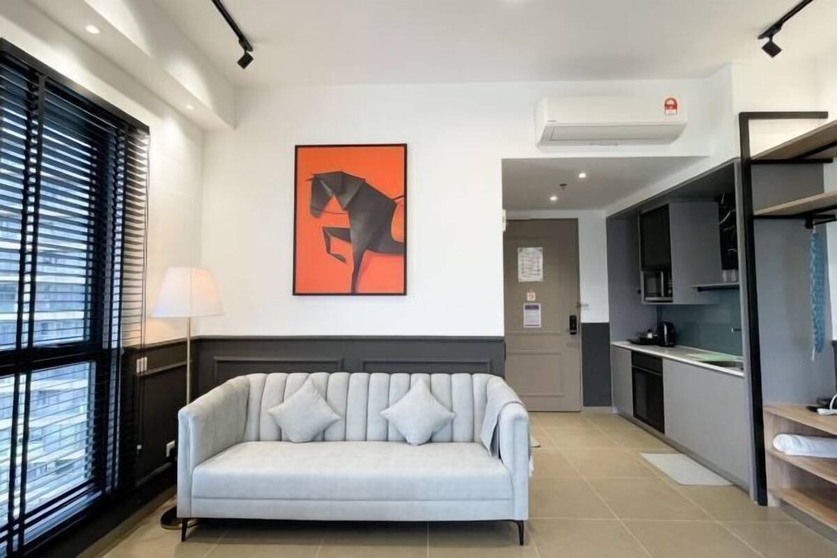 Una habitación de hotel moderna con sofá, un cuadro, una ventana con persianas y una kitchenette.