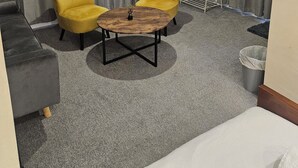 Ένα δωμάτιο ξενοδοχείου με έναν γκρι καναπέ, δύο κίτρινες πολυθρόνες, ένα ξύλινο τραπεζάκι σαλονιού και ένα κρεβάτι με λευκά σεντόνια.