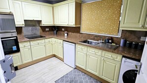 Μια κουζίνα με ανοιχτόχρωμα πράσινα ντουλάπια, πάγκο από γρανίτη, εντοιχισμένο φούρνο, πλυντήριο πιάτων και πυροσβεστήρα.