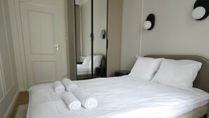 Ein ordentlich bezogenes Bett mit weißen Leinentüchern und aufgerollten Handtüchern, ein Nachttisch und ein Spiegel an der Wand.