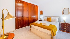Quarto com um guarda-roupa de madeira grande, cama com manta amarela mostarda, mesa de cabeceira com abajur e espelho na parede.