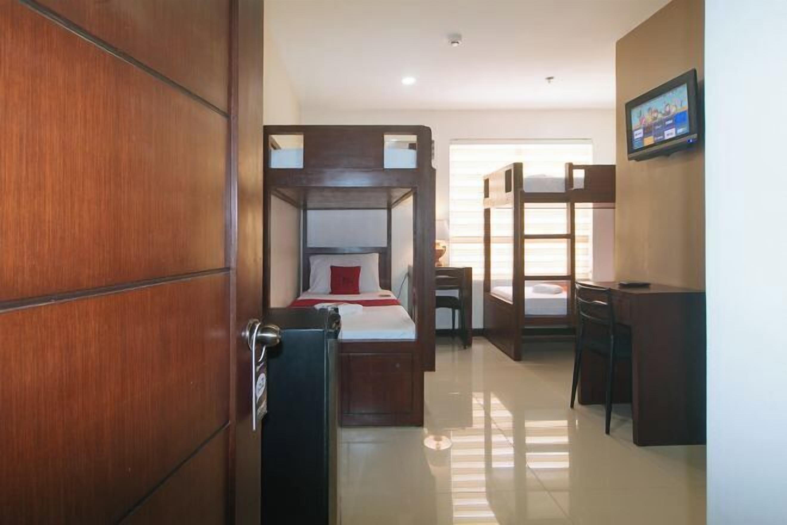 A hotel room with a wooden wardrobe, a bed with a red pillow, a desk, and a television on the wall.