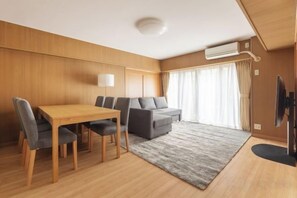 A modern hotel room with a wooden floor, a dining table with chairs, a grey sofa, a flat-screen TV, and a large window with curtains. - HAN HOTEL Shinjuku (Tokyo)
