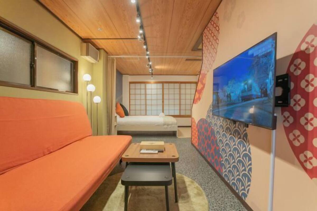 Quarto de hotel com teto de madeira, um sofá, uma cama, uma mesinha e uma televisão fixada na parede.
