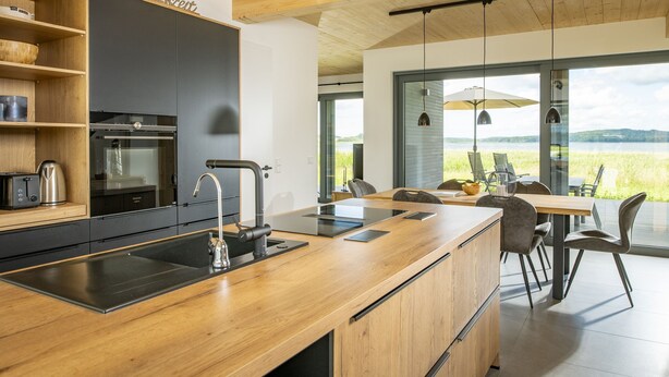 A modern kitchen with a wooden countertop, black cabinets, and a view of a lake through large windows. - House "LUV" on the lake in Streu (Bergen auf Rügen)