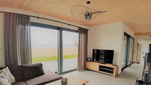 A modern living room with a sofa, a coffee table, and a TV on a wooden stand. Large windows offer a view of the outdoors. - House "LUV" on the lake in Streu (Bergen auf Rügen)