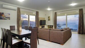 Un salon moderne avec un grand canapé, une table à manger et des chaises. Un téléviseur à écran plat est fixé au mur, et de grandes fenêtres offrent une vue sur la mer et les montagnes.