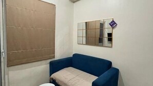 A room with a blue sofa, a small round table, a beige throw blanket, a window with a beige blind, and a mirror on the wall. - DLP Rooms (Central Jakarta)