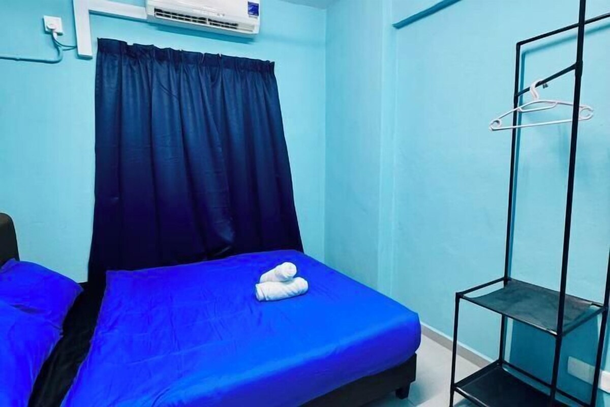 A small, single-bed room with a blue bedspread, a black metal shelf unit, and a white air conditioning unit mounted on the wall.
