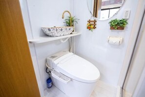 Een kleine badkamer met toilet, een plankje met een kom en een plant, een spiegel en een klein raam.