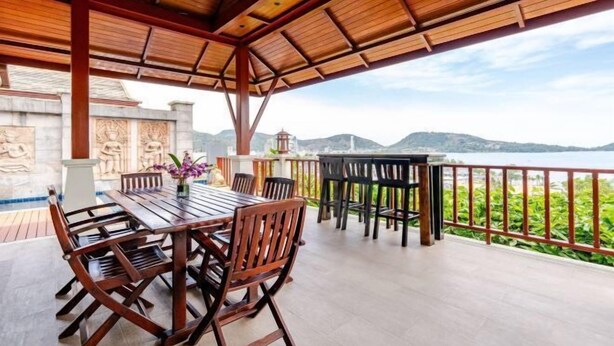 Un área de comedor al aire libre techada, con mesas y sillas de madera, vista al mar y una pared de piedra con relieves.