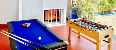 A pool table with a cue stick, a foosball table, and a dartboard in a room with a view of greenery outside.