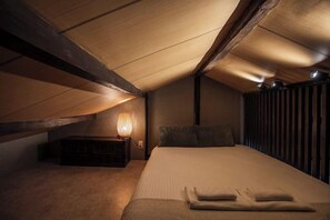 Un dormitorio con techo inclinado, una cama con almohadas, una mesita de noche con lámpara y una ventana con persianas.