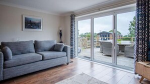 A modern living room with a grey sofa, a patterned rug, and a large sliding glass door leading to an outdoor patio area. - Ridgeway Meadow Bungalow - 3 Bedroom - Manorbier (Tenby)