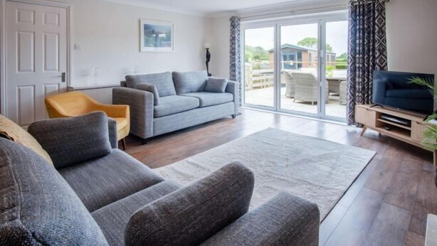 A modern living room with a grey sofa, a yellow armchair, a flat-screen TV, and a large sliding glass door leading to an outdoor patio. - Ridgeway Meadow Bungalow - 3 Bedroom - Manorbier (Tenby)